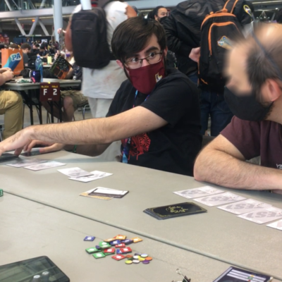 A set of cards are spread out across a tabletop. In the background, a young man is gesturing to some of the cards and looking at an older man who has a similar set of cards in front of him. Both are wearing masks and are of pale European complexion. A large crowd are visible in the background.